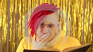 Young man with red dreadlocks reading book. Stylish hipster in eyeglasses surprised opening his mouth from plot book.