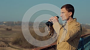 Young man with red hair in in khaki coat looking through binoculars from the bridge.