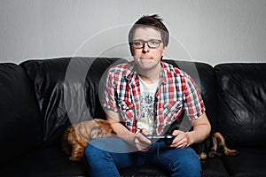 The young man in the red shirt and glasses playing video games with a joystick sitting on a black leather couch with two dogs. He