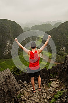 Young man in red sports shirt raises his arms victoriously after reaching the top of a viewpoint in Tam Coc