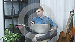 Young man relaxing in cozy chair at modern apartment, using phone and laptop. Male sitting at home office with laptop computer. Bu