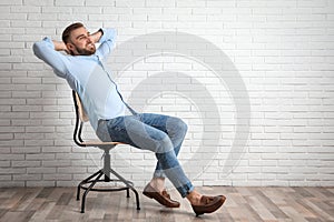 Young man relaxing in office chair near white brick wall indoors, space for text