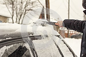 A young man removes snow from the roof and car windows with a brush. Heavy snowfall and cyclone