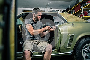 Young man removing interior trim panels from an old vintage car from the 60s or 70s in his home garage. Tools are seen around