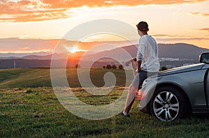 Young man resting during long solo auto trip sitting on his luxury car bonnet and enjoying the sunset sky colors. Traveling by car