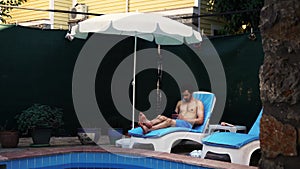 Young man is resting next to the swimming pool on a lounge chair and using smartphone in his hands