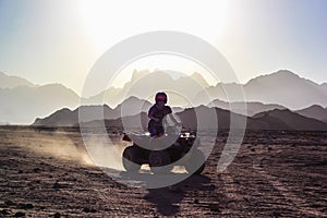 Young man rides an ATV on the desert over background of mountains at sunset
