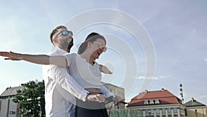 Young man rides his girlfriend on an electric scooter around the city square. Happy girl laughs and spreads her arms to