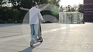 Young man rides his girlfriend on an electric scooter around the city square. Happy girl laughs and spreads her arms to