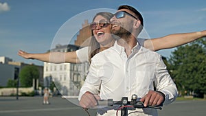 Young man rides his girlfriend on an electric scooter around the city square. Happy girl laughs and spreads her arms to