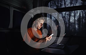 Young man rides a night train with a laptop at the table and uses a smartphone with a serious face