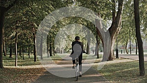 Young man riding a bike in city park wearing business suit and helmet, back view