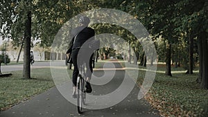 Young man riding a bike in city park wearing business suit and helmet, back view