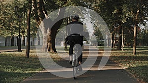 Young man riding a bike in city park wearing business suit and helmet, back view