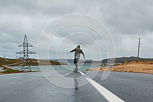 Young man riding a longboard on empty winding mountain road in summer, rear view.