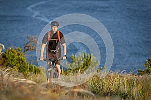 Young man riding mountain bike on the green meadow above the blue river in the countryside.