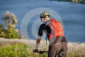 Young man riding mountain bike on the green meadow above the blue river in the countryside.