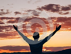 Young man rising up the hands and greeting the stunning sunset orange-pink sky colors over the mountain landscape back view. New