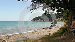 Young man rocking girl in bikini on rope swing under tree against calm azure sea