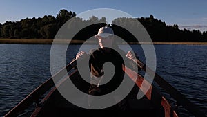 Young man is rowing with wooden oars on wooden boat on a lake during sunny day