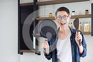 Young man`s standing holding the pan and ladle. He`s looking at the camera and smiling in his kitchen for prepared to cook