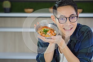 Young man`s wearing glasses sitting looking at the camera, smiling and showing the fried egg with colorful toppings served on a