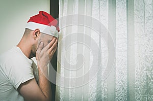 Young man with Santa Claus hat cover his face with his hands and cry near window feeling lonely and sad for New Year and Christmas