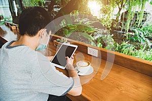 A young man sat on a wooden chair typing on a digital tablet in a coffee shop with a clear glass wall, outside with a garden and g