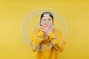 Young man with a scared shocked face covers his mouth with his hands and looks at the camera on a yellow background