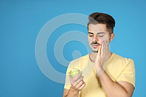 Young man with sensitive teeth and apple on color background.