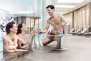 Young man serving with champagne two women at the swimming pool