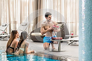 Young man serving with champagne two women at the swimming pool