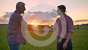 Young man shaking hands with old farmer, standing on wheat field and watching beautiful sunset in background