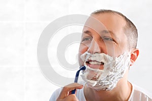 A young man shaves with a disposable razor in the bathroom while looking in the mirror. Health, skin care