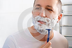 A young man shaves with a disposable razor in front of a mirror. Health, skin care