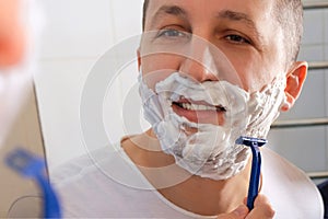A young man shaves with a razor in front of a mirror.