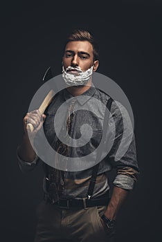 Young man with shaving cream on his face holding an axe and looking