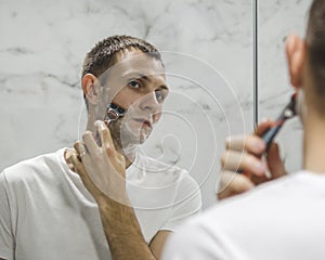 A young man is shaving his beard near the mirror
