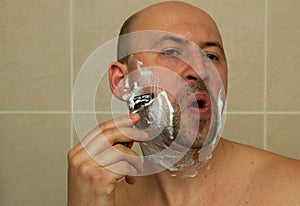 Young man shaving his beard with razor reflected