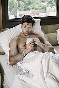 Young man shirtless on his bed with a coffee or tea cup