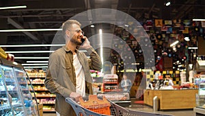 Young man with shopping trolely in a supermarket talks on the phone, consults with his wife about the shopping list