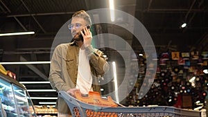 Young man with shopping trolely in a supermarket talks on the phone, consults with his wife about the shopping list