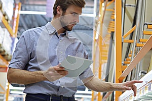 Young man shopping or working in a hardware warehouse standing checking supplies on his tablet