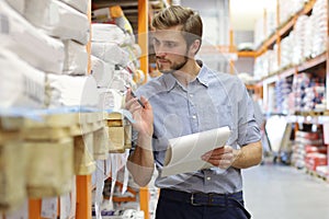 Young man shopping or working in a hardware warehouse standing checking supplies on his tablet