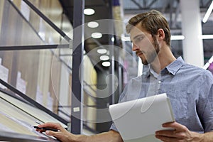 Young man shopping or working in a hardware warehouse standing checking supplies on his tablet