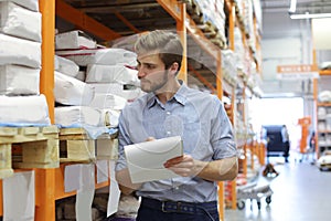 Young man shopping or working in a hardware warehouse standing checking supplies on his tablet