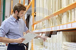 Young man shopping or working in a hardware warehouse standing checking supplies on his tablet