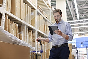 Young man shopping or working in a hardware warehouse standing checking supplies on his tablet