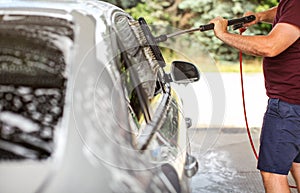 Young man in shorts and t shirt washing his car in self serve carwash, cleaning side windows with brush