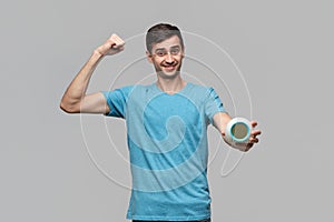 Young man showing his bicep holding alarm isolated over grey background. Time to go for workout